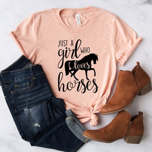 May include: A peach-colored t-shirt with the text "Just a girl who loves horses" in black lettering. The shirt is tied in a knot at the bottom and is paired with blue jeans and brown ankle boots.