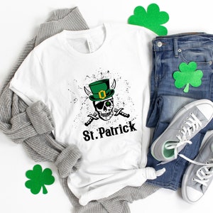 May include: White t-shirt with a green leprechaun skull graphic and the text "St. Patrick". The shirt is laid out on a white surface with a gray sweater, blue jeans, and green shamrock decorations.