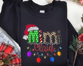 Könnte beinhalten: Schwarzer Pullover mit einem weihnachtlichen Design mit dem Text "Mimi Claus" in Grün, Rot und Weiß mit einer Weihnachtsmütze und Leopardenmuster-Akzenten. Das Design enthält auch eine Lichterkette.