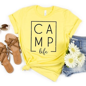 May include: Yellow t-shirt with black text that reads "CAMP life" inside a black square. The shirt is styled with brown sandals, denim shorts, and white flowers.