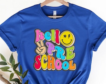 May include: A royal blue t-shirt with the text "hello preschool" in colorful retro letters. A yellow smiley face with stars is above the word "pre".