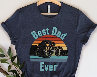 May include: A navy blue t-shirt with a vintage-style graphic that says "Best Dad Ever" with two fists bumping. The graphic features a sunset with orange, yellow, red, and blue colors.
