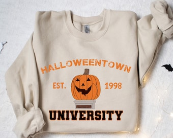 Może przedstawiać: Beżowa bluza z pomarańczową grafiką uśmiechniętej dyni na cokole, z napisami "Halloweentown University" i "Est. 1998". Czarne wycinanki nietoperzy i łańcuchy świetlne zdobią tło.