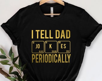 Può includere: Maglietta nera con testo dorato che recita "Dico barzellette a papà periodicamente". Il testo è stilizzato per assomigliare a una tavola periodica con gli elementi "Jo", "K" ed "Es" evidenziati.