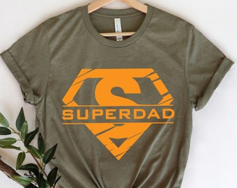 May include: Olive green t-shirt with an orange graphic of a stylized "S" inside a diamond shape, with the word "SUPERDAD" below. The shirt is displayed on a wooden hanger against a white background, with a sprig of greenery.