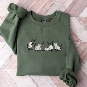 May include: An olive green sweatshirt featuring an embroidered design of cats dressed as ghosts with bats. The design is in black and white. The sweatshirt has long sleeves with ribbed cuffs and a crew neck.