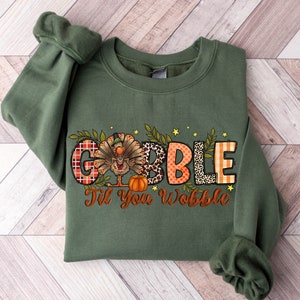 May include: Green sweatshirt with a Thanksgiving-themed design. The design features a turkey with a pumpkin and the words "Gobble Til You Wobble".