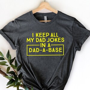 May include: A dark gray t-shirt with a yellow graphic that reads "I keep all my dad jokes in a dad-a-base".