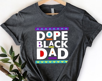 May include: A dark grey t-shirt with a white graphic that says "Dope Black Dad" in colorful block letters. The graphic is surrounded by a colorful geometric pattern.