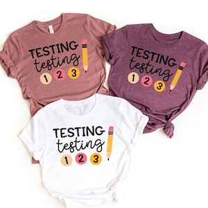 May include: Three t-shirts in mauve, purple, and white, each featuring the words "TESTING testing" in black, with the numbers 1, 2, and 3 in circles, and a yellow pencil graphic. The shirts are made of soft material.
