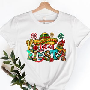 Puede incluir: Camiseta blanca con un dise&ntilde;o gr&aacute;fico colorido que dice "Let's Fiesta". El dise&ntilde;o incluye un sombrero, maracas y otros elementos festivos.