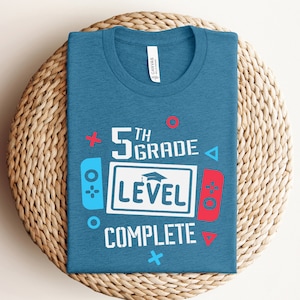 May include: A blue t-shirt with a graphic design that says "5th Grade Level Complete" with a graduation cap and a video game controller design.