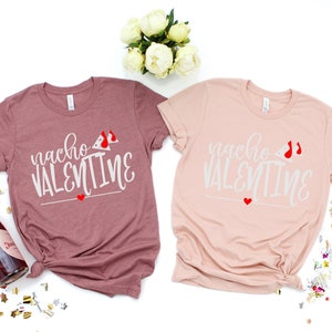May include: Two t-shirts with the text "nacho valentine" and a red heart and a red and white nacho cheese design. The shirts are pink and mauve.