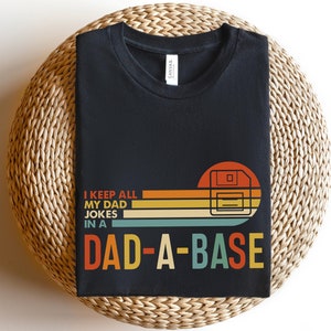 May include: Black t-shirt with a retro-style graphic of a floppy disk and the text "I keep all my dad jokes in a Dad-A-Base".
