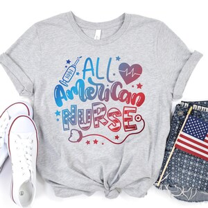 K&ouml;nnte beinhalten: Ein graues T-Shirt mit dem Text "All American Nurse" in Rot, Wei&szlig; und Blau. Der Text ist in einer stilisierten Schriftart gehalten und enth&auml;lt ein Herz mit einem Herzschlag-Symbol, eine Spritze und Sterne.
