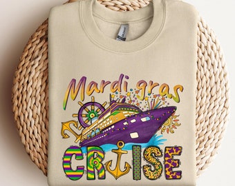 Op de afbeelding: Een beige sweatshirt met een kleurrijk Mardi Gras-cruiseontwerp. Het ontwerp bevat een paars en gouden cruiseschip met een stuurwiel, anker en de woorden "Mardi Gras Cruise".