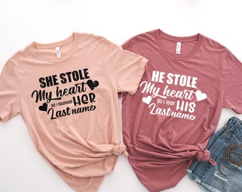 May include: Two t-shirts with a matching design. The pink shirt says "She stole my heart so I changed her last name" with a black heart and the text in black. The maroon shirt says "He stole my heart so I took his last name" with a white heart and the text in white.