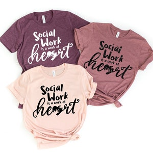 May include: Three t-shirts with the text "Social Work is a work of heart" in black lettering with a handprint heart. The shirts are in different shades of pink and purple.
