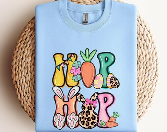 May include: Light blue sweatshirt with a graphic design of the word "HOP" in colorful letters with Easter-themed decorations, including carrots, eggs, and a bunny.
