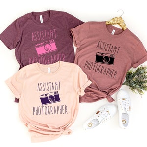 May include: Three t-shirts with the text "Assistant Photographer" and a vintage camera graphic. The shirts are in shades of pink and maroon.
