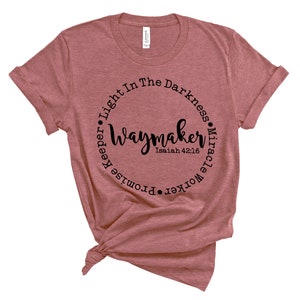 May include: A dusty rose t-shirt with a round black graphic. The graphic reads "Waymaker" in cursive, surrounded by the words "Light In The Darkness, Miracle Worker, Promise Keeper." The text "Isaiah 42:16" is below "Waymaker."