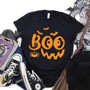 Puede incluir: Camiseta negra con un dise&ntilde;o de cara de calabaza naranja y la palabra "BOO" en una fuente estilizada. El dise&ntilde;o incluye una ara&ntilde;a y murci&eacute;lagos.