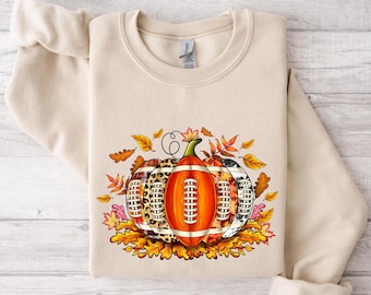 May include: A beige sweatshirt with a graphic of a football-shaped pumpkin with fall leaves. The footballs are patterned with plaid, leopard print, and camouflage.