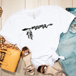 May include: White t-shirt with a black arrow and feathers design and the word "mama" printed on it. The shirt is laid out on a wooden surface with a pair of gold sandals, a yellow handbag, and a pair of blue jeans.