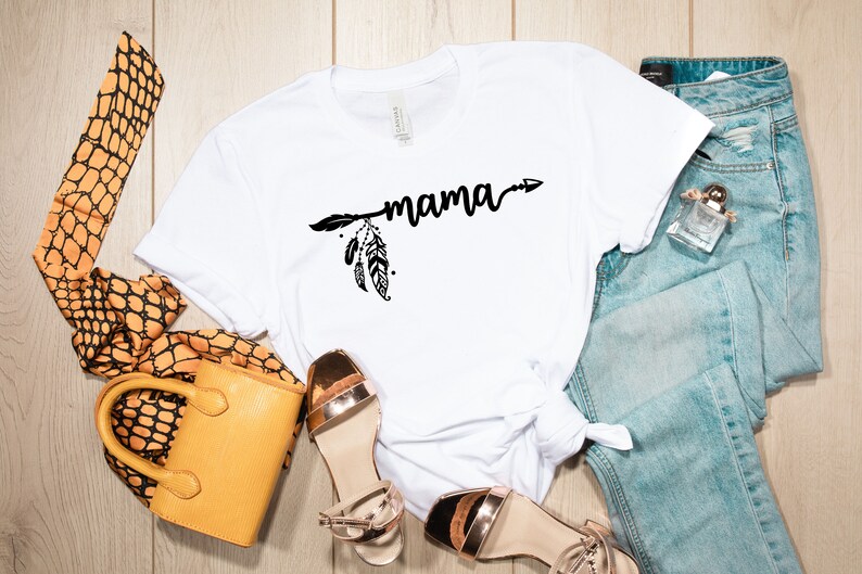 May include: White t-shirt with a black arrow and feathers design and the word "mama" printed on it. The shirt is laid out on a wooden surface with a pair of gold sandals, a yellow handbag, and a pair of blue jeans.