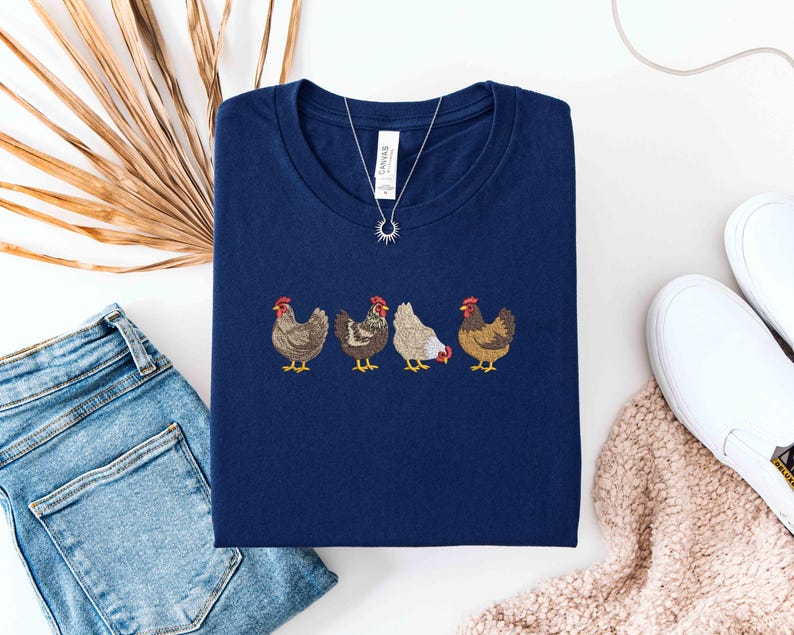 May include: Navy blue t-shirt featuring an embroidered design of four chickens in various colors. The chickens have red combs and yellow feet. A silver necklace with a sunburst pendant is on the shirt. The shirt is paired with blue jeans and white sneakers.