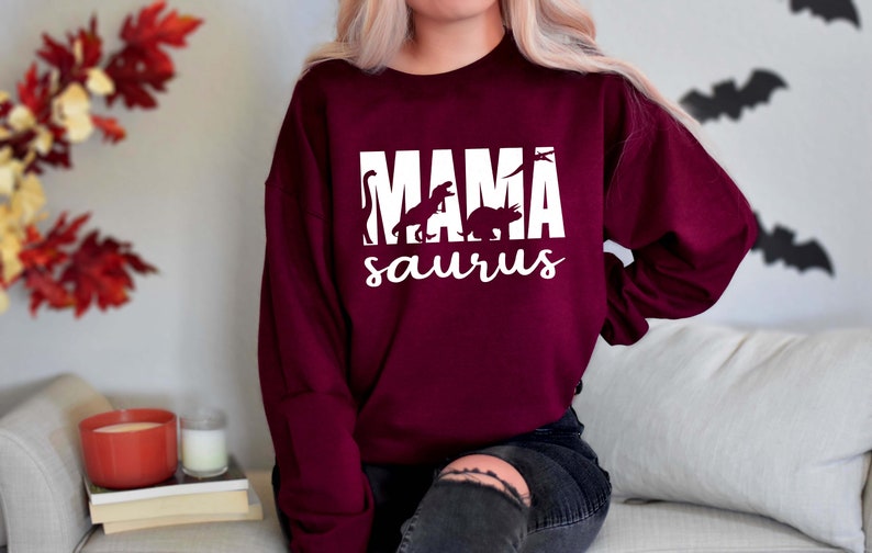 May include: A maroon sweatshirt with the text "MAMA saurus" in white. The text is surrounded by silhouettes of dinosaurs.