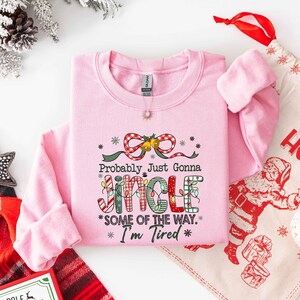 Pu&ograve; includere: Una felpa rosa chiaro con la scritta "Probably Just Gonna Jingle Some of the Way. I'm Tired" in un design festivo. Il design include un fiocco, luci natalizie e fiocchi di neve. Sullo sfondo &egrave; visibile un sacchetto regalo natalizio.