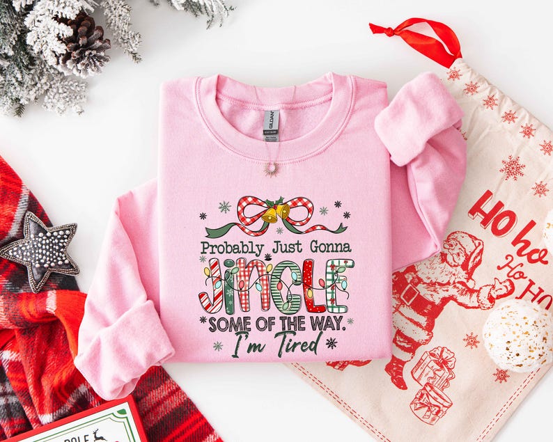 Pu&ograve; includere: Una felpa rosa chiaro con la scritta "Probably Just Gonna Jingle Some of the Way. I'm Tired" in un design festivo. Il design include un fiocco, luci natalizie e fiocchi di neve. Sullo sfondo &egrave; visibile un sacchetto regalo natalizio.