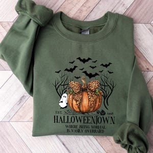 May include: Olive green sweatshirt featuring a Halloween-themed graphic. The design includes a pumpkin with a leopard print bow, bats, a ghost, and the text "Halloweentown". The phrase "Where being normal is vastly overrated" is also included.