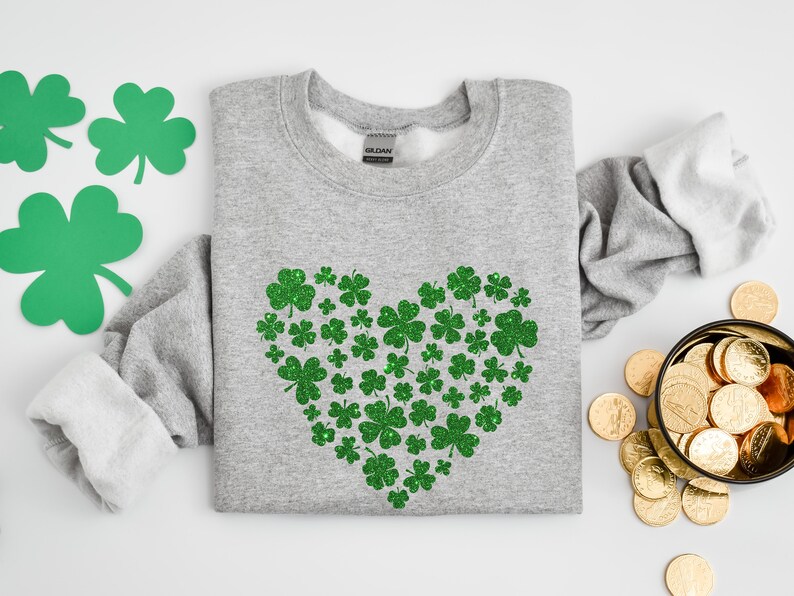 May include: A grey sweatshirt with a green glitter shamrock heart design. The shamrocks are arranged in a heart shape on the front of the sweatshirt.