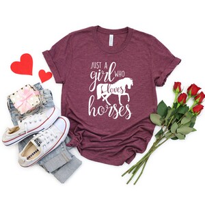 May include: A maroon t-shirt with the text "Just a girl who loves horses" in white lettering. The shirt features a white silhouette of a horse. The shirt is displayed on a white background with a pair of white sneakers, a pair of jeans, a gift box, and a bouquet of red roses.