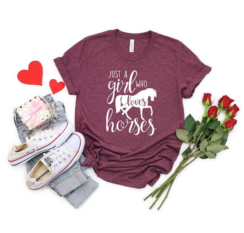 May include: A maroon t-shirt with the text "Just a girl who loves horses" in white lettering. The shirt features a white silhouette of a horse. The shirt is displayed on a white background with a pair of white sneakers, a pair of jeans, a gift box, and a bouquet of red roses.