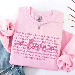 May include: A light pink sweatshirt with the word "love" in a cursive font and the text of 1 Corinthians 13:4-7 printed in a darker pink. A silver necklace with a small pendant rests on the shirt.