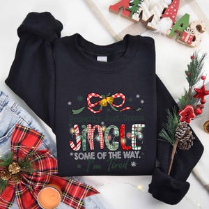Pu&ograve; includere: Felpa nera con la scritta "Probably Just Gonna Jingle Some of the Way. I'm Tired" in un design natalizio. Il design include un fiocco, campane e luci fatate. La felpa &egrave; mostrata con jeans e decorazioni festive.
