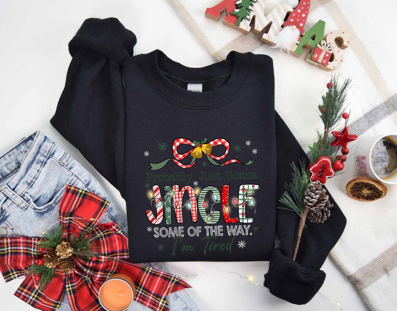 Pu&ograve; includere: Felpa nera con la scritta "Probably Just Gonna Jingle Some of the Way. I'm Tired" in un design natalizio. Il design include un fiocco, campane e luci fatate. La felpa &egrave; mostrata con jeans e decorazioni festive.