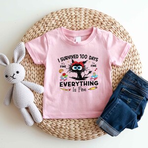 May include: Pink short-sleeved t-shirt with the text "I Survived 100 Days, It's Fine, Everything Is Fine" and a cartoon cat graphic. The shirt is paired with blue denim jeans and a gray stuffed bunny.