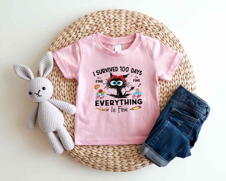 May include: Pink short-sleeved t-shirt with the text "I Survived 100 Days, It's Fine, Everything Is Fine" and a cartoon cat graphic. The shirt is paired with blue denim jeans and a gray stuffed bunny.