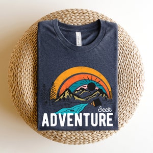 May include: Dark blue t-shirt with a colorful graphic of a mountain landscape, rainbow, and the text "Seek Adventure" in white. The design includes a sun, trees, and a river. The shirt is folded on a woven surface.