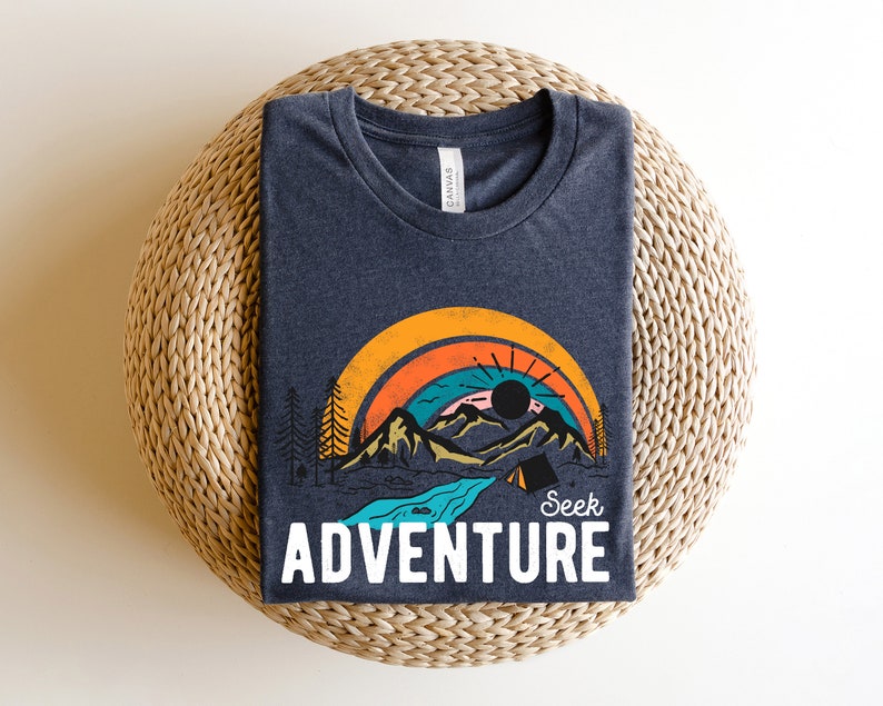 May include: Dark blue t-shirt with a colorful graphic of a mountain landscape, rainbow, and the text "Seek Adventure" in white. The design includes a sun, trees, and a river. The shirt is folded on a woven surface.