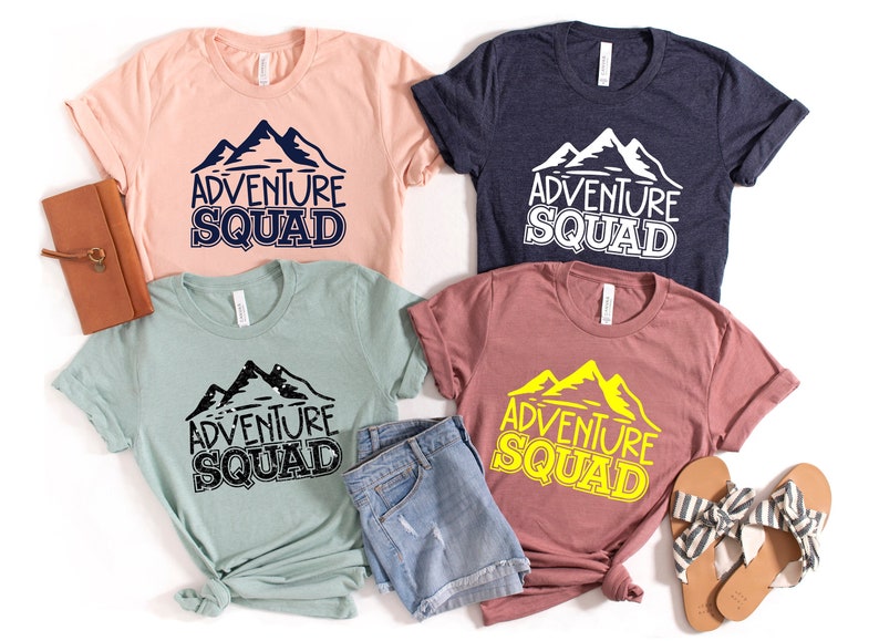 Puede incluir: Cuatro camisetas con el texto "Adventure Squad" y un gr&aacute;fico de monta&ntilde;a. Las camisetas son de diferentes colores: rosa, azul, verde y marr&oacute;n. El gr&aacute;fico de monta&ntilde;a es de diferentes colores en cada camiseta: negro, blanco, negro y amarillo.