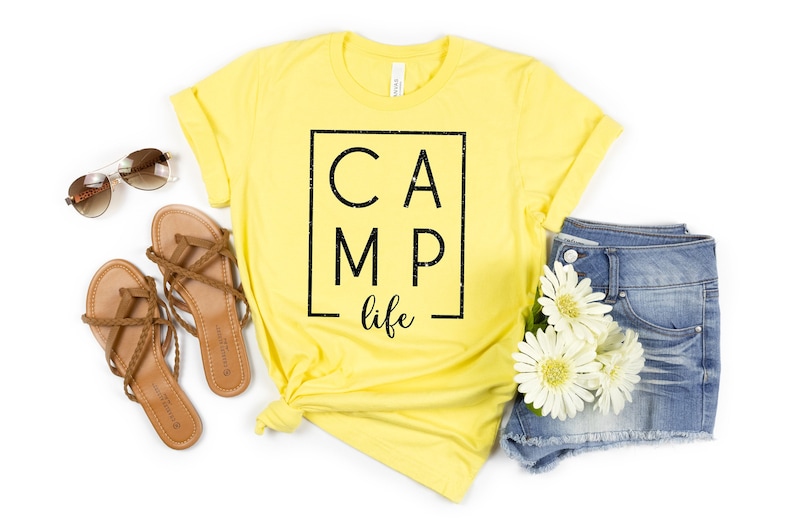 May include: Yellow t-shirt with black text that reads "CAMP life" inside a black square. The shirt is styled with brown sandals, denim shorts, and white flowers.