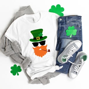 May include: A white t-shirt with a green leprechaun face graphic. The leprechaun is wearing a green hat with a gold buckle, sunglasses, and a red beard with a pipe. The shirt is laid out on a white surface with a gray sweater, blue jeans, and green shamrock cutouts.