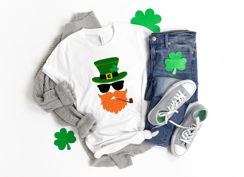 May include: A white t-shirt with a green leprechaun face graphic. The leprechaun is wearing a green hat with a gold buckle, sunglasses, and a red beard with a pipe. The shirt is laid out on a white surface with a gray sweater, blue jeans, and green shamrock cutouts.