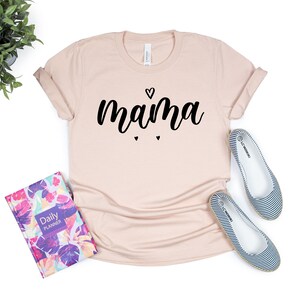 May include: A light pink t-shirt with the word "mama" written in black cursive lettering with a small heart above the word and two smaller hearts below the word.