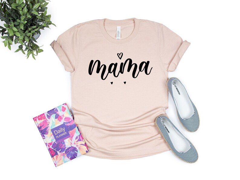 May include: A light pink t-shirt with the word "mama" written in black cursive lettering with a small heart above the word and two smaller hearts below the word.